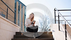 Young blonde woman works with a tablet sitting on the stairs outside and drinking coffee