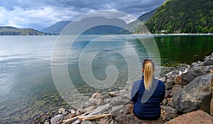 Young blondie woman sitting on the edge of a wooden dock on Harrison Lake and watching the calm before the storm