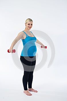 Young blondy pregnant woman in blue tank top does exercises with small red fitness dumbbells, white background.