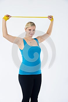 Young blondy pregnant woman in blue tank top does exercises with yellow fitness rubber band, white background.