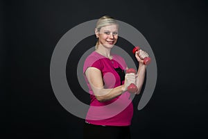 Young blondy pregnant woman in pink t-short with cat does exercises with red fitness dumbbells, black background.