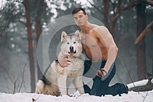 Young bodybuilder with bare torso embraces dog Malamute at walk in winter forest.