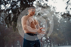 Young bodybuilder with bare torso wipes his body by snow in forest in winter.