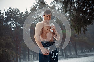 Young bodybuilder with bare torso wipes his body by snow in forest in winter.