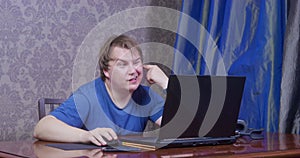 Young bored man in blue t-shirt with a silly look picks at his ear and scratches it, sits at table in front of laptop