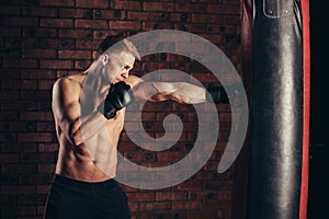 A young boxer in black gloves with a naked torso work out strikes on punching bag.