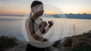 Young boxer girl posing by the sea over huge cliffs at sunset. She wears tights and sports bra