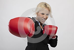 Young boxing businesswoman punching towards camera wearing red boxing gloves, selective focus, closeup, light background