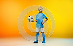 Young boy as a soccer or football player in sportwear practicing on gradient yellow studio background in neon light