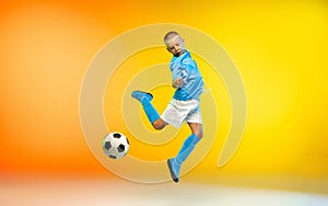 Young boy as a soccer or football player in sportwear practicing on gradient yellow studio background in neon light