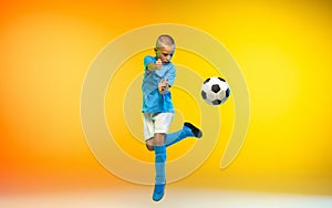 Young boy as a soccer or football player in sportwear practicing on gradient yellow studio background in neon light