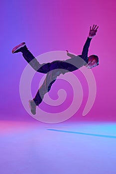 Young boy in black attire, street dancer performing tricks in mid-air in mixed neon light against vibrant gradient