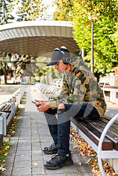 Young boy with black cap sitting on bench in park and using smarthphone