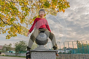 Young boy in casual clothes sits on a decorative fence with marble ball near an autumn