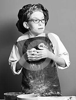 Young boy cute cook chef in uniform and hat on stained face flour with glasses standing near table with colorful bawls