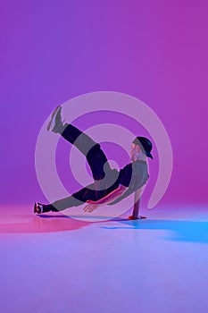 Young boy, dancer in motion performing energetic street dance in mixed neon light against vibrant gradient background.