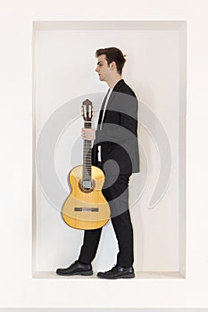 A young boy in an elegant suit stands in a niche in the wall. He is holding his guitar with one hand