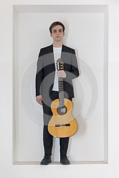 A young boy in an elegant suit stands in a niche in the wall. With one hand he is holding his guitar