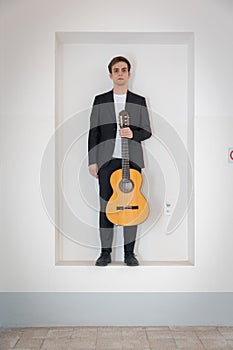 A young boy in an elegant suit stands in a niche in the wall. With one hand he is holding his guitar