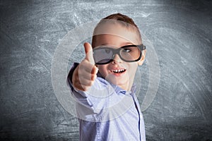 Young boy with eyeglasses in front of a school chalkboard showing thumbs up. Eye examination, education and learning concept