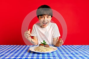 Young boy feeling excited, sitting at table with blue checkered tablecloth and plate of spaghetti and meatballs against