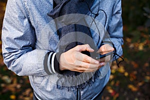 Young boy in gray jacket with gray scarf holds and uses smartphone with headphones outside over autumn background. Technology