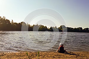 A young boy in a hat sits on the beach and plays with the sand next to the lake.