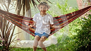 Young boy in a hat sits and sways in a garden hammock, capturing carefree childhood memories and the relaxation of a