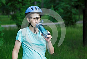 Young boy in helmet and green t shirt cyclist drinks water from bottle in the park. Smiling cute Boy on bicycle in the forest