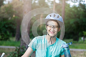 Young boy in helmet and green t shirt cyclist drinks water from bottle in the park. Smiling cute Boy on bicycle in the forest