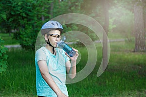 Young boy in helmet and green t shirt cyclist drinks water from bottle in the park. Smiling cute Boy on bicycle in the forest