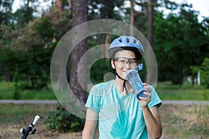 Young boy in helmet and green t shirt cyclist drinks water from bottle in the park. Smiling cute Boy on bicycle in the forest