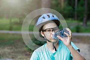 Young boy in helmet and green t shirt cyclist drinks water from bottle in the park. Smiling cute Boy on bicycle in the forest