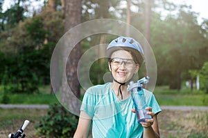Young boy in helmet and green t shirt cyclist drinks water from bottle in the park. Smiling cute Boy on bicycle in the forest