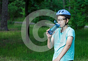 Young boy in helmet and green t shirt cyclist drinks water from bottle in the park. Smiling cute Boy on bicycle in the forest