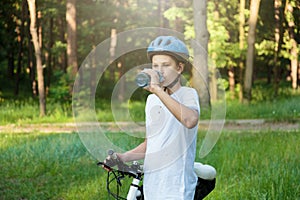 Young boy in helmet and white t shirt cyclist drinks water from bottle in the park. Smiling cute Boy on bicycle in the forest