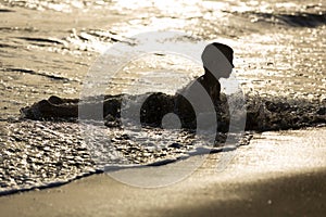 Young boy lays in water at the sandy beach, sunset