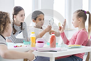 A young boy looking through a magnifying glass and a girl holding a slice of cucumber while other kids are watching during