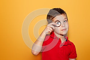A young boy looking through a magnifying glass in red t-shirt, on pure Yellow background. Searching. Space for text
