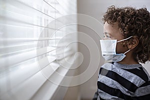 Young boy looking out the window wearing a protective facemark while seeking protection from COVID-19, or the novel coronavirus