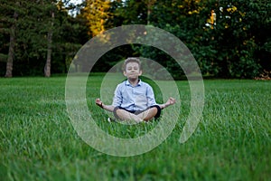 Young boy is meditating in the lotus position on a background of green meadows at the park.