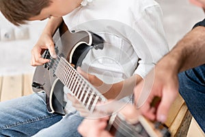 Young boy moves his hand along guitar strings while his father helps boy to take chords