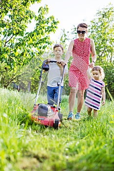 Young boy mowing grass with lawn mower, his mother and sister encouraging him