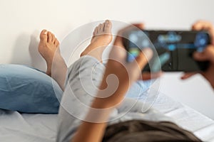 Young boy in pajamas lying on bed and playing video game on mobile phone