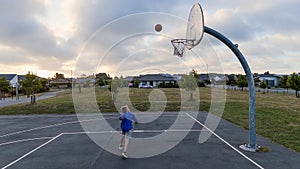 A young boy plays basketball under the cloudy sky in the park court