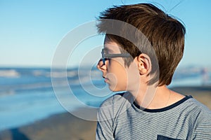Young boy posing at the summer beach. Cute calm spectacled 12 years old boy at seaside, lokking at sea.