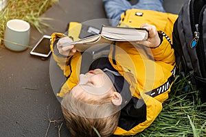 A young boy reads a book in a clearing. He is lying on a tourist rug on the green grass, a cup is nearby, and a telephone is lying