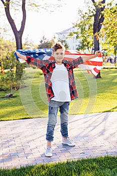 A young boy in a red plaid shirt and white T-shirt happily standing in the park with the stretched USA flag of America and smiling