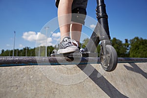 Young boy riding on a kick scooter in a concrete skatepark. Unrecognizable kid standing on a skate ramp top on a push-scooter.