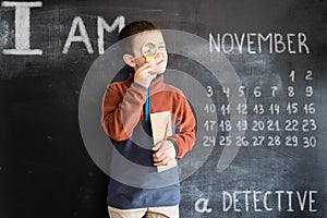 Young boy`s standing with magnifying glass and notebook in his hands near blackboard. Young detective. Creative design